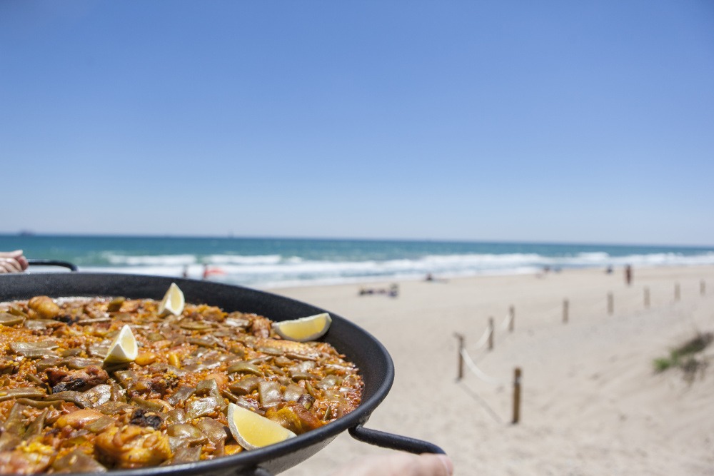 En stor paellapanna med citronklyftor hålls upp mot en solig strandbakgrund.