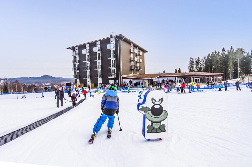Ett barn åker skidor i snön framför ett stort hotell, med en tecknad vargfigur på en skylt i förgrunden och skogklädda berg i bakgrunden.