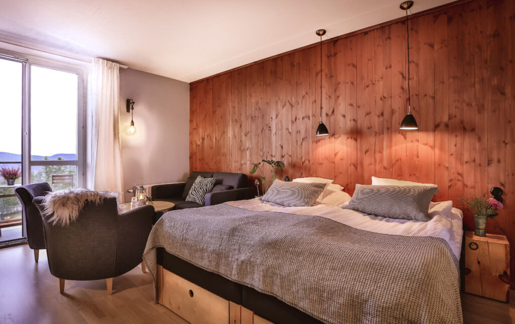 Ett mysigt hotellrum med bruna träpanelväggar, en stor säng med grått överkast, mörka fåtöljer och en balkong med utsikt mot fjällen.