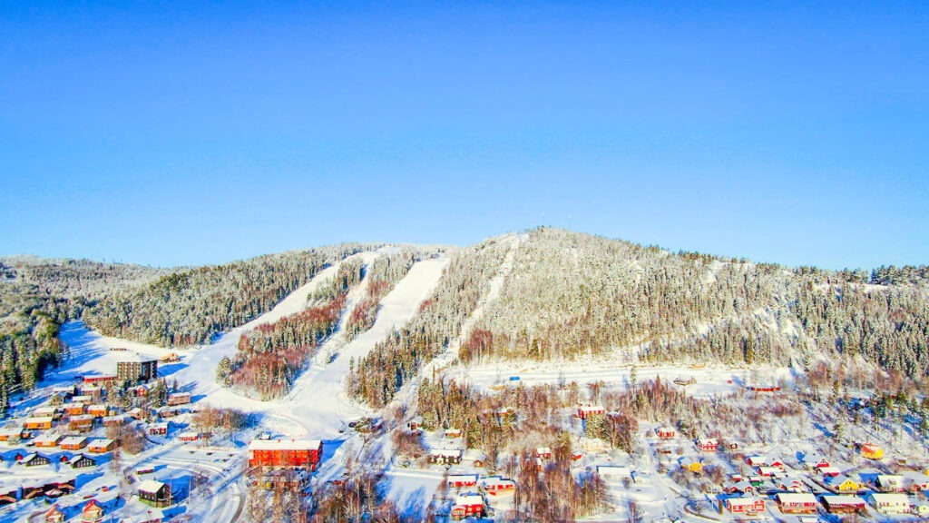 Ett flygfoto över en skidort med snötäckta backar på ett berg, omgivet av vinterklädda skogar och färgglada byggnader i dalen nedanför.