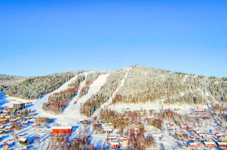 Ett flygfoto över en skidort med snötäckta backar på ett berg, omgivet av vinterklädda skogar och färgglada byggnader i dalen nedanför.