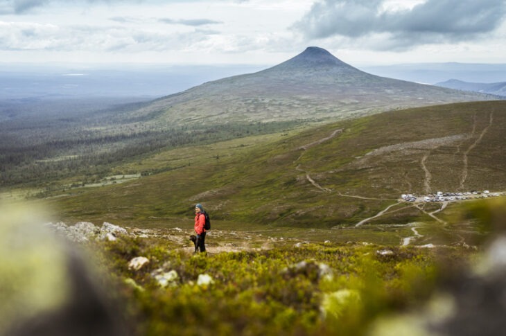 En person i röd jacka står på ett fjäll med en stor topp i bakgrunden, omgiven av gröna sluttningar och skog.