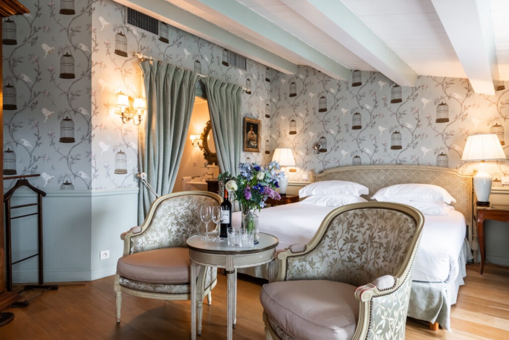 Ett hotellrum med tapet med fågelburar, två fåtöljer, en säng med vita lakan och en vas med blommor.