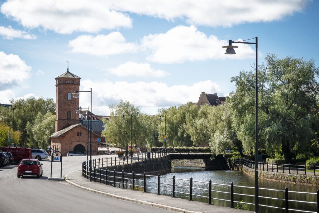 En stadsgata med en tegelbyggnad med torn vid en kanal, omgiven av gröna träd och en klarblå himmel med vita moln.