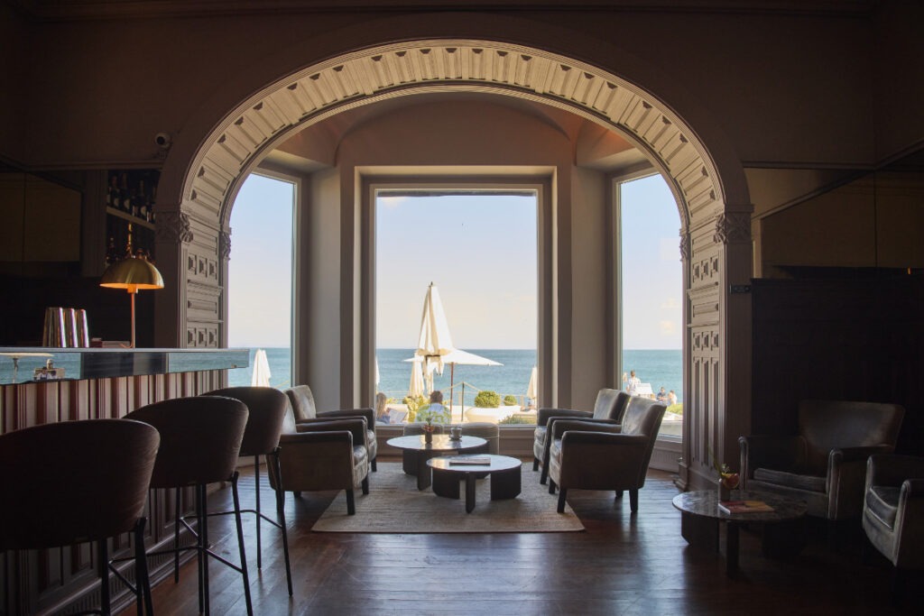 En hotellounge med mörka trägolv och skinnfåtöljer, en stor rundad dörröppning mot havet, bardisk med mässingslampa och vita parasoller på terrassen utanför.