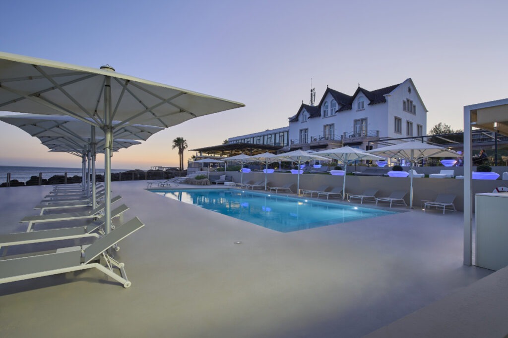 En upplyst pool vid skymning med blått ljus i vattnet, omgiven av vita solstolar och parasoller, med ett vitt hotellhus och havet i bakgrunden.