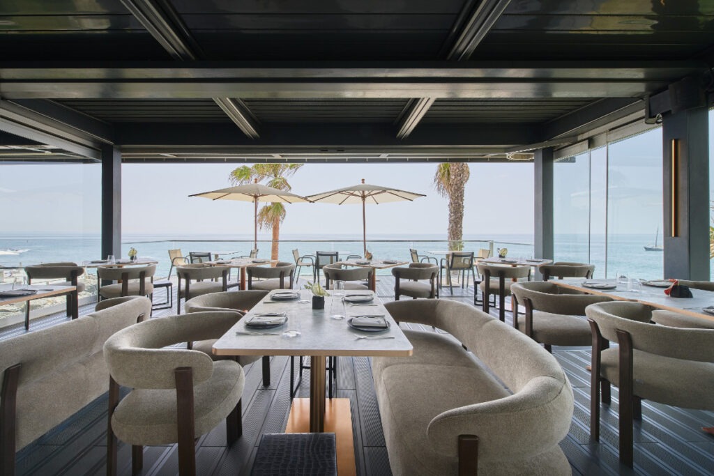 En modern restaurang med stora fönster mot havet, gråa stolar och soffor vid dukade bord, palmträd och en segelbåt synlig på vattnet utanför.