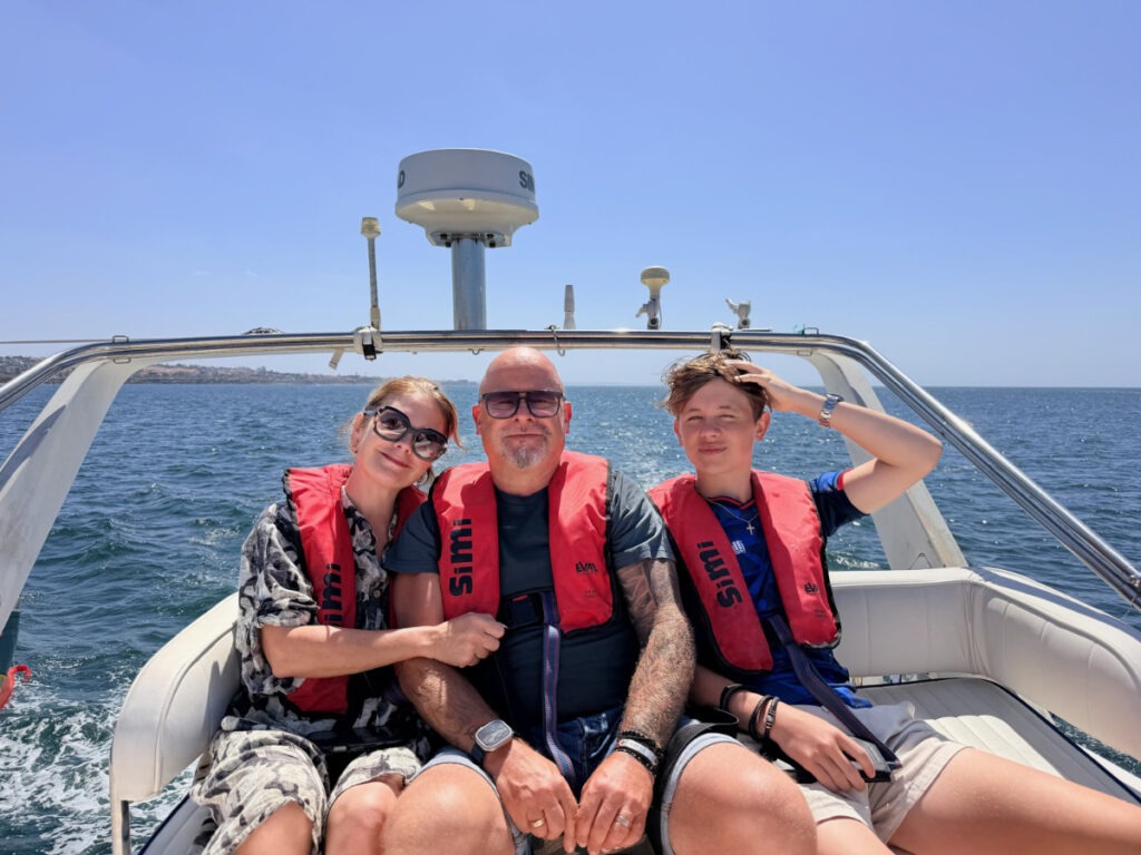 Tre personer i röda flytvästar sitter i en båt på havet, med en klarblå himmel och vatten i bakgrunden.