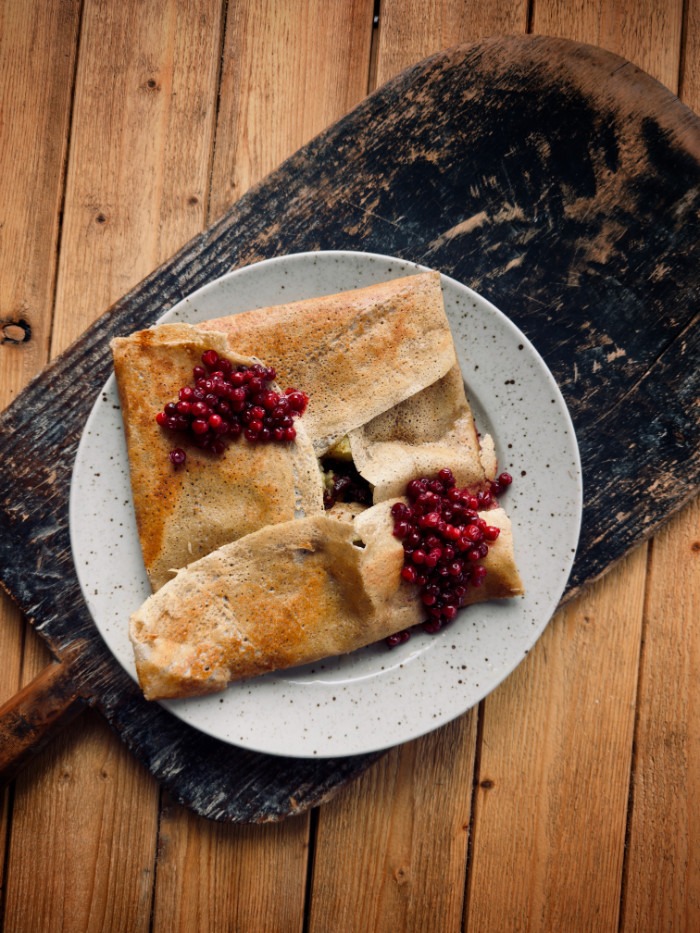 En tallrik med två ihopvikta pannkakor toppade med lingon, serverade på en sliten träskärbräda mot en träbakgrund.