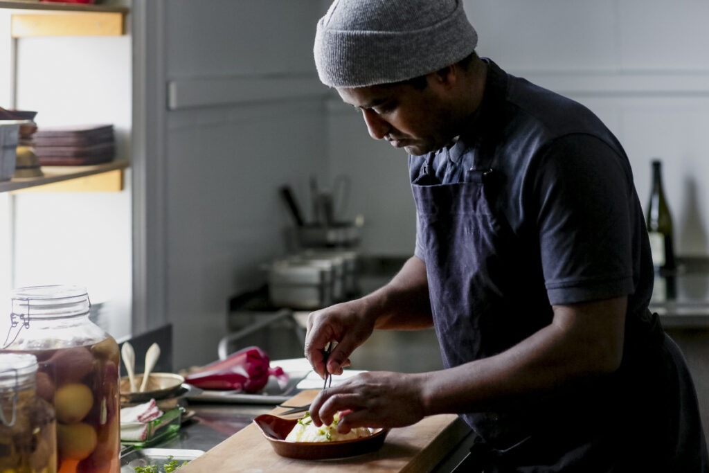 En kock i grå mössa och mörkt förkläde garnerar en rätt i ett restaurangkök, med glasburkar av inlagda grönsaker på disken.