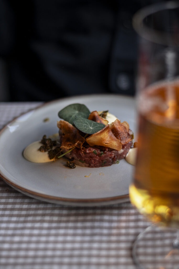 En tallrik med råbiff toppad med krispiga potatischips och gröna blad, med ett glas öl i förgrunden på en rutig duk.