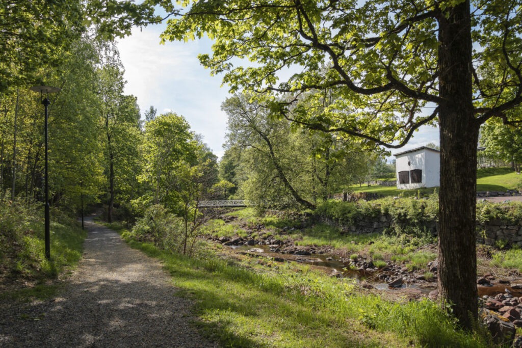 En grusväg löper längs en bäck med stenar i en grönskande skog, med en vit byggnad synlig på en kulle i bakgrunden.