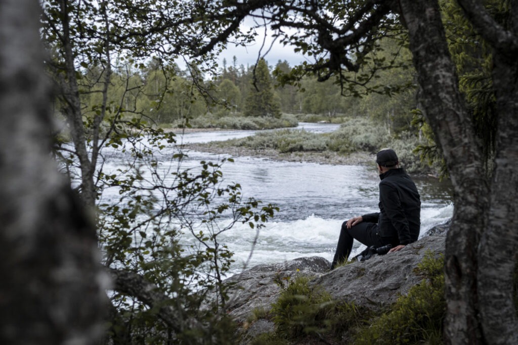 En man sitter på en sten vid en forsande älv i en skog, omgiven av träd och grönska.