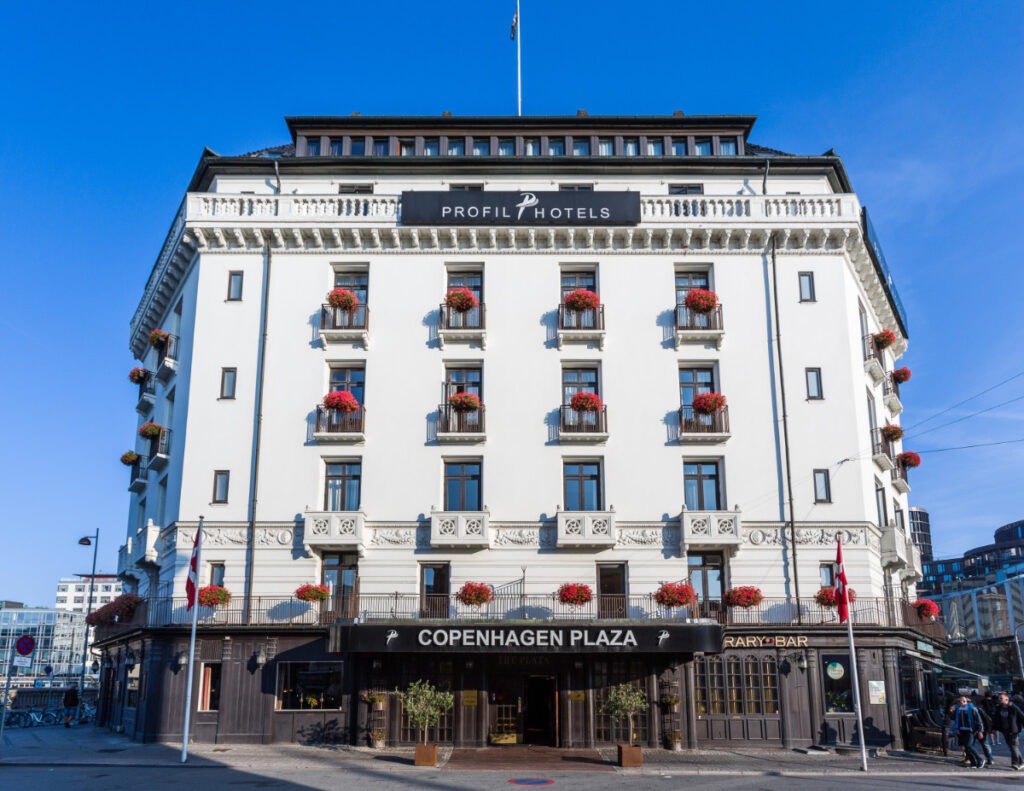 Profil Hotels Copenhagen Plaza, ett vitt stenhus med röda blommor på balkongerna och mörk bottenvåning.