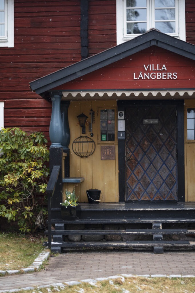 Entrén till Villa Långbers, ett rött trähus med gul dörr och svarta detaljer, med snöflingor som faller.