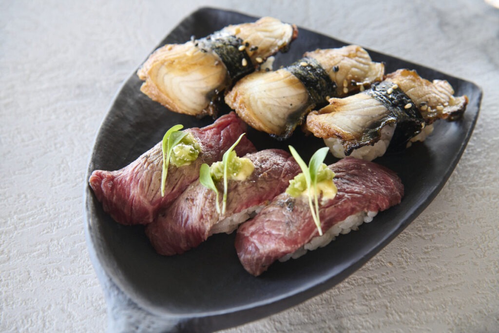 Ett svart fat med sex nigiri-sushibitar, tre med grillat nötkött och wasabi samt tre med ål och sesamfrön, på en ljus texturerad yta.
