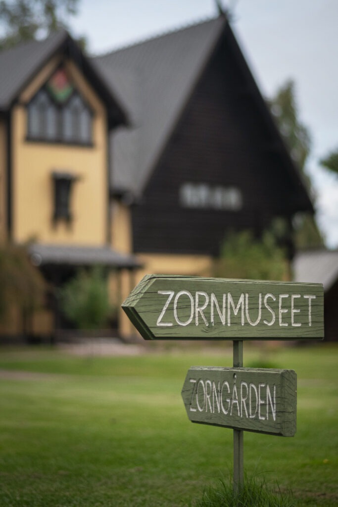 En träskylt visar vägen till "ZORNMUSEET" och "ZORNGÅRDEN", med ett gult hus med svart tak i bakgrunden.