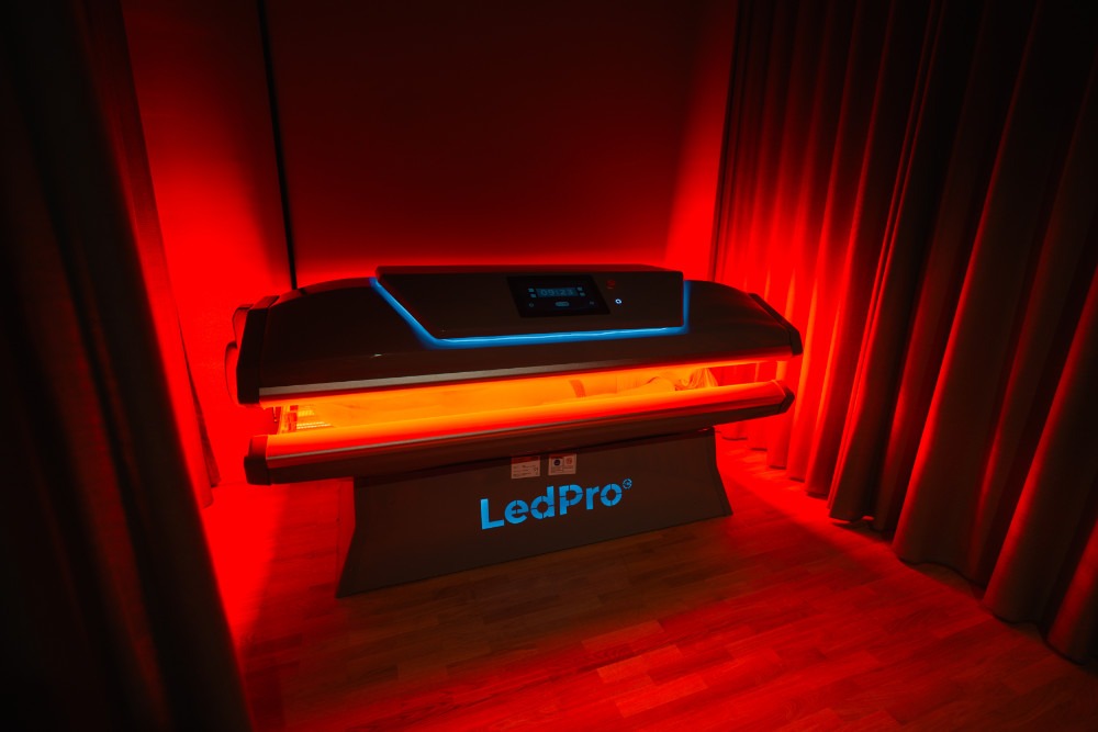 Rött LED-solarium med skärm och mörka förhängen i rum med trägolv.