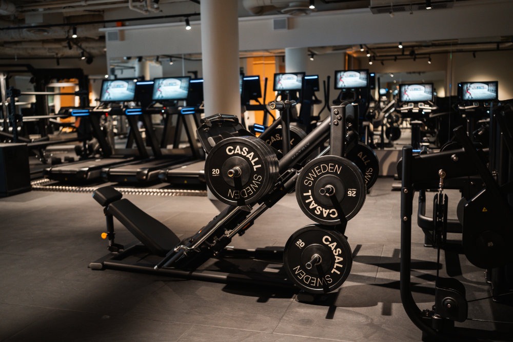 Benpressmaskin med Casall-viktskivor på mörkt gym, löpband med skärmar i bakgrunden.